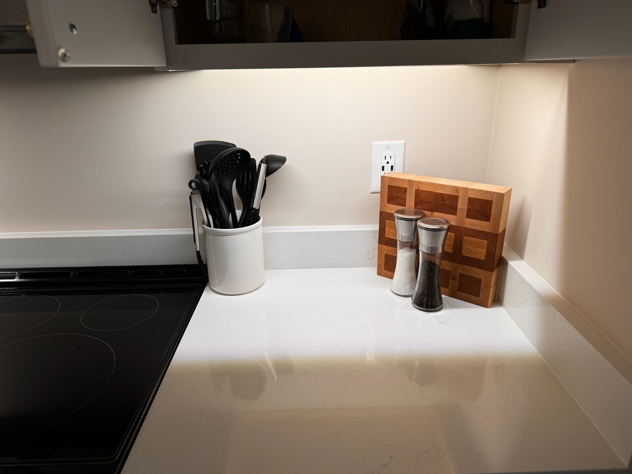 Eine Küchenarbeitsplatte mit einer weißen Tasse, in der Besteck liegt, einem Holzregal für Gewürze und einem Herd.
