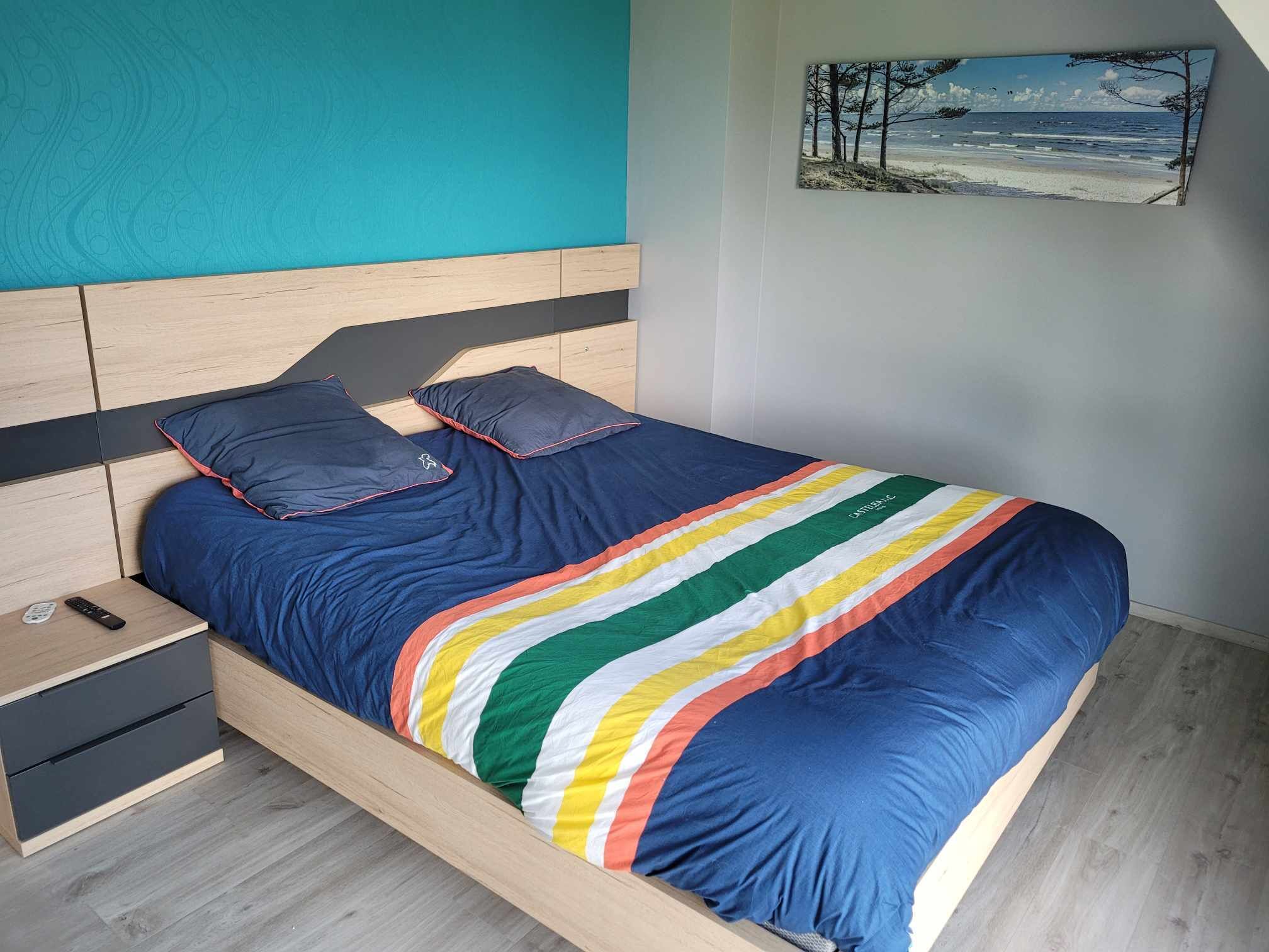 A bedroom with a wooden headboard, a bed with a striped blanket, two pillows, a nightstand with a remote control, and a painting on the wall.