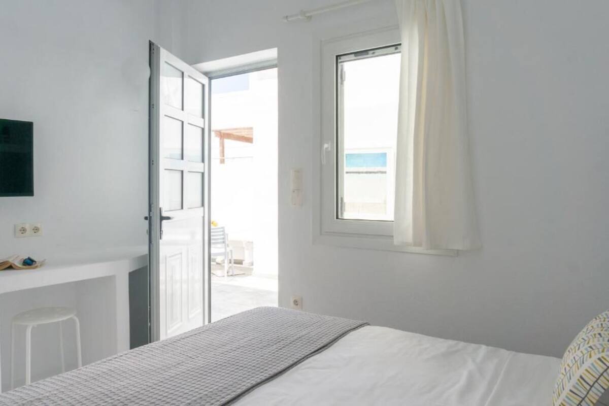 A bedroom with a bed, a window with curtains, a door leading to an outdoor area, and a small stool.