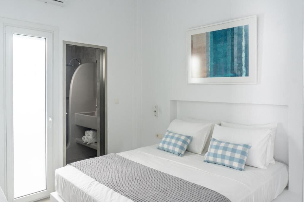 Un dormitorio con una cama, dos almohadas, una almohada a cuadros, una obra de arte enmarcada y un baño con ducha y toallero.