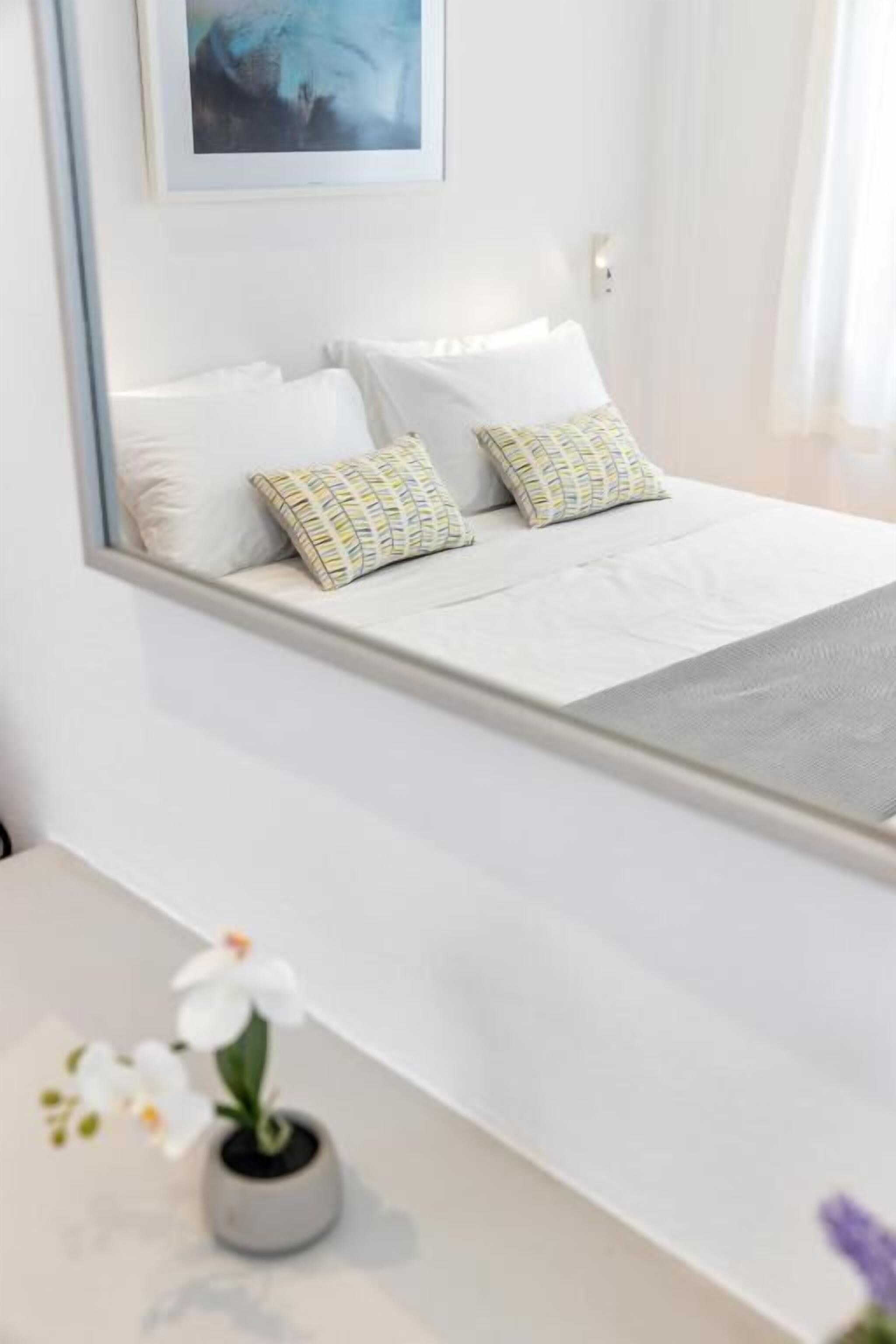 Un lit bien fait, avec des draps blancs et des coussins à motifs, une petite plante en pot sur la table de chevet et une œuvre d’art encadrée au-dessus du lit.