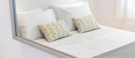 Un lit bien fait, avec des draps blancs et des coussins à motifs, une petite plante en pot sur la table de chevet et une œuvre d’art encadrée au-dessus du lit.