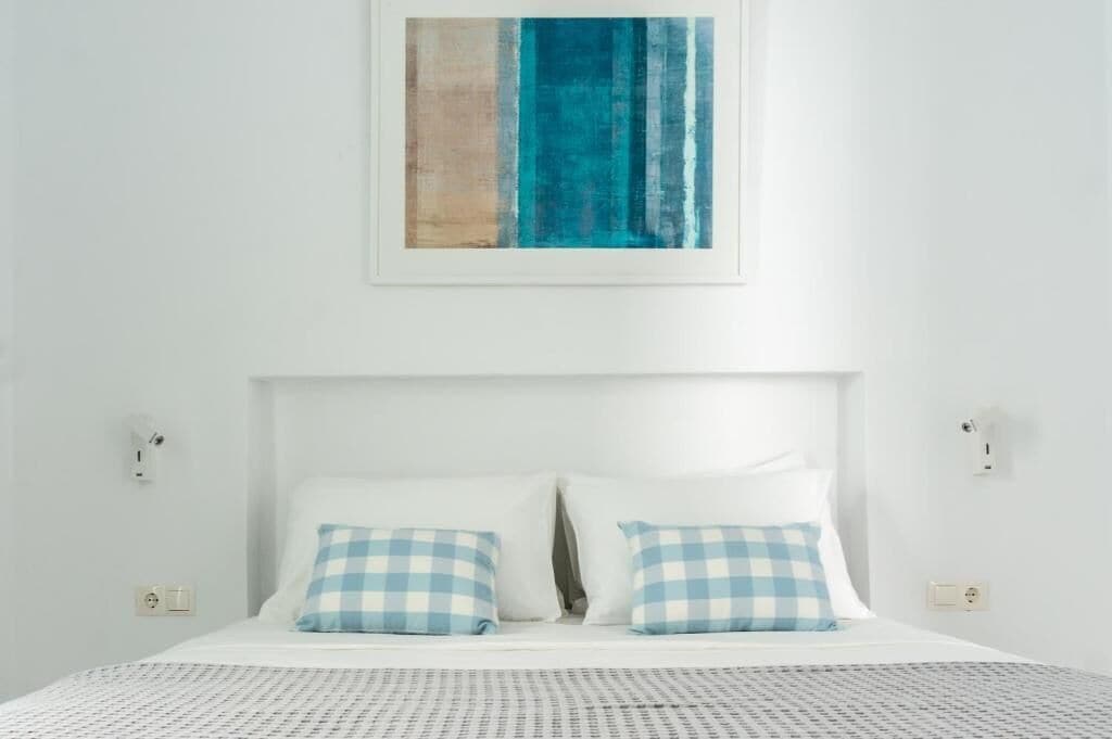 A neatly made bed with checkered pillows, a plain white wall, and a framed abstract painting above it.