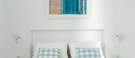 A neatly made bed with checkered pillows, a plain white wall, and a framed abstract painting above it.