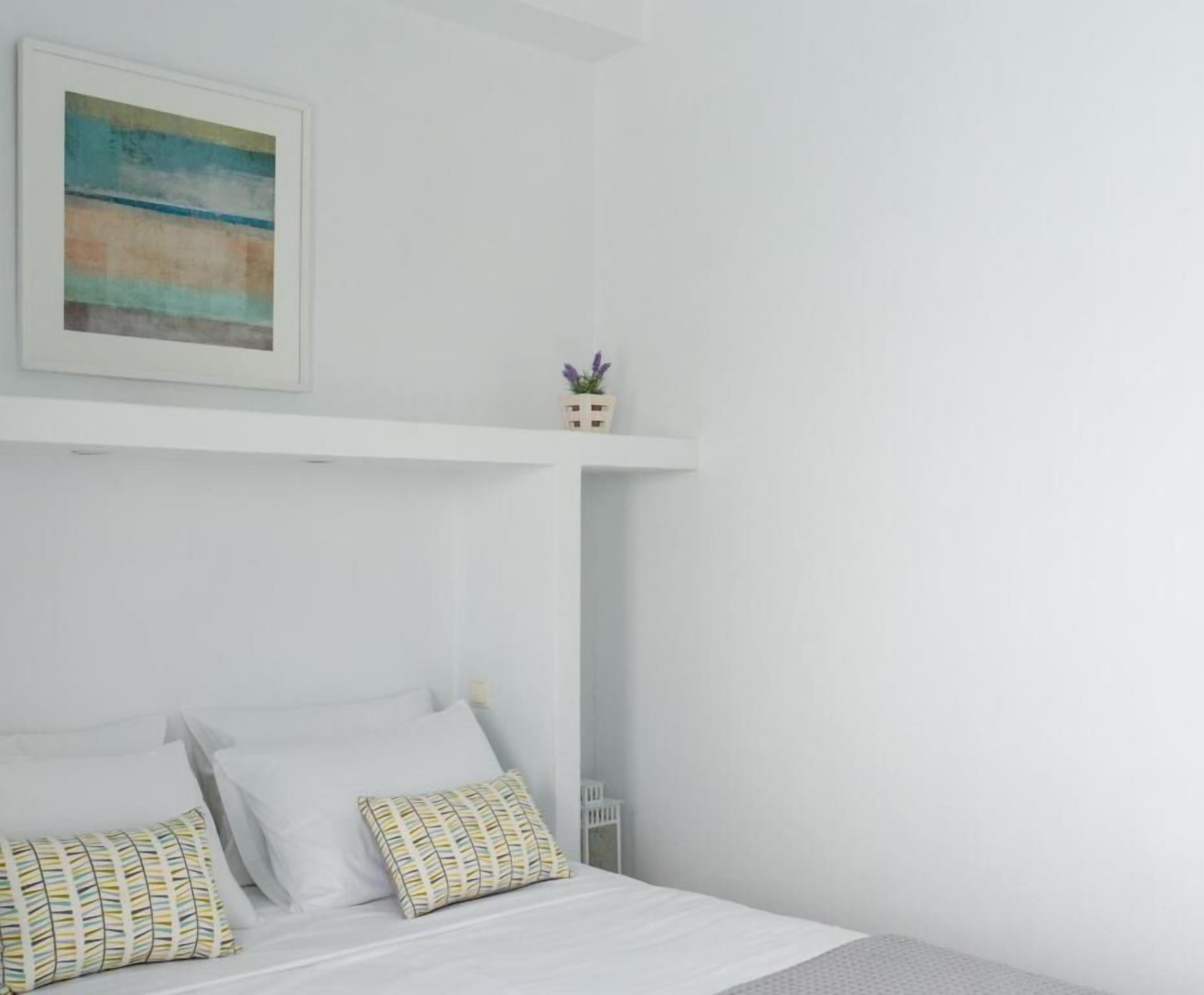 Un dormitorio con una cama, almohadas, una mesita de noche, un estante con una planta y un cuadro en la pared.