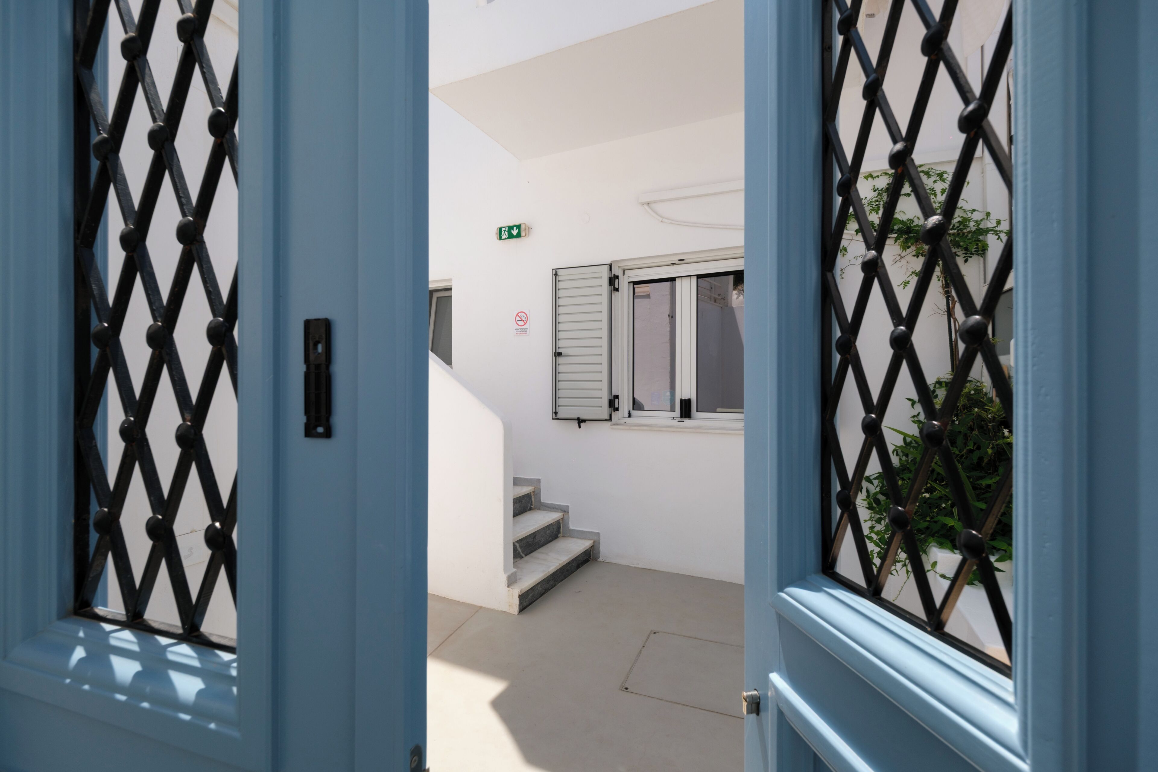 Una vista a través d'una porta blava amb una reixa de metall, que dona a una escala i una finestra amb persianes.