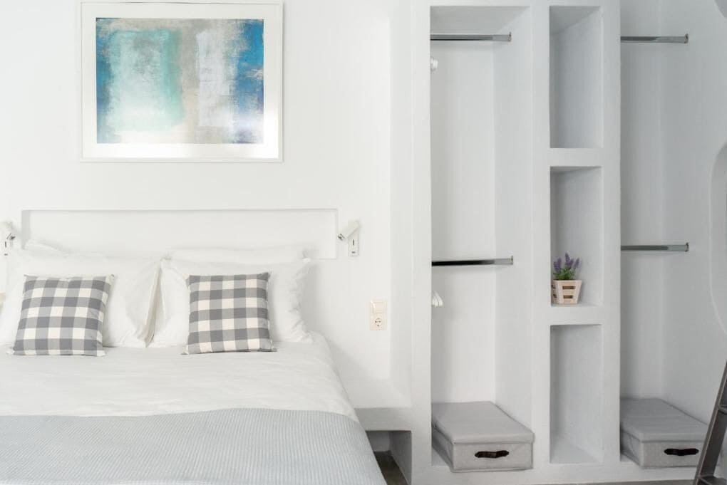 A bedroom with a bed, checkered pillows, a framed abstract painting, and a white wardrobe with shelves and drawers.