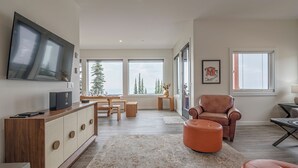 Una sala de estar moderna con un televisor grande, muebles de madera y vistas a árboles a través de amplios ventanales.
