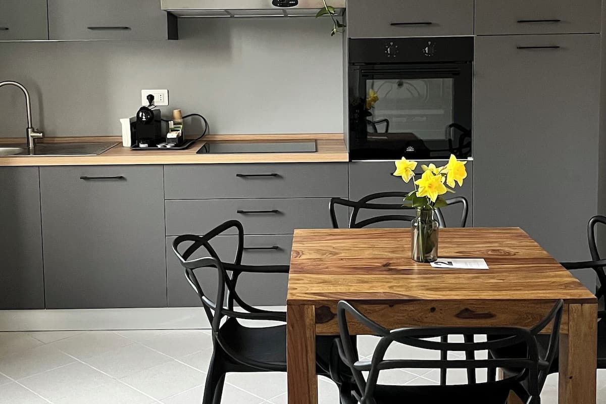 Una cocina moderna con armarios grises, una mesa de comedor de madera y sillas negras. Hay una cafetera en la encimera y una planta en maceta en el estante.