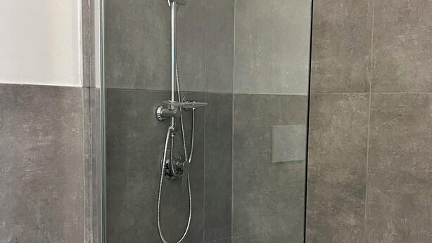 Un baño moderno con ducha acristalada, lavamanos blanco y baldosas grises.