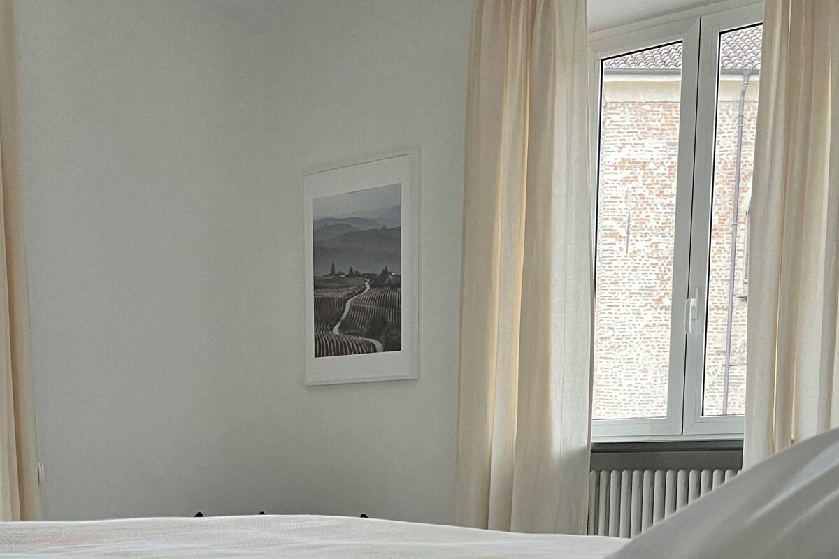 Un dormitorio con una cama, un radiador, una ventana con cortinas e imagen enmarcada en la pared.