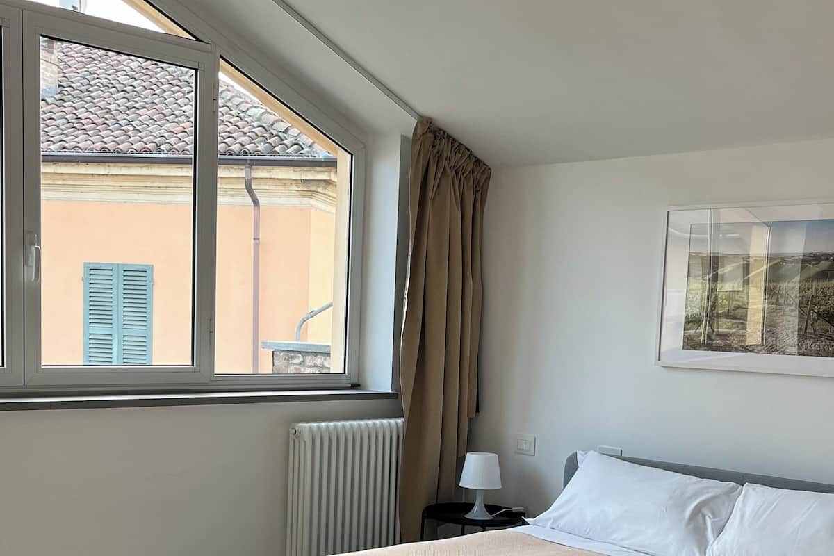 Un dormitorio con cama, ventana, radiador, mesita de noche, lámpara y un cuadro en la pared.