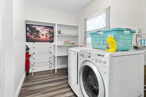 A laundry room with a washing machine, a drying rack, a shelf with toys, a basket, and a bottle of detergent. - Perfect Location for Sports and Concert Venues! - La Cabina (Tampa)