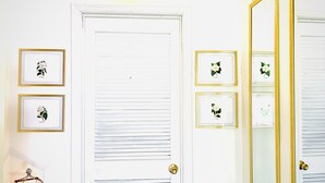 A white door with horizontal blinds, three framed botanical prints on the wall, a small table with a decorative lamp, and a large mirror with a gold frame. - The Gardenia Suites in The Heart of Spring Hill-A (Spring Hill)