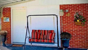 A covered porch with a swing seat, a door, and a brick wall with a flower basket. - The Gardenia Suites in The Heart of Spring Hill-A (Spring Hill)