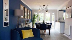 A modern living room with a blue sofa, a dining area with a table and chairs, and a kitchen in the background. - Beautiful seaside retreat close to North Devon's white sandy beaches! (Wrafton)