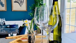 A dining table with a bottle of wine, two wine glasses, and a wooden board with crackers and a small vase with flowers. - Beautiful seaside retreat close to North Devon's white sandy beaches! (Wrafton)