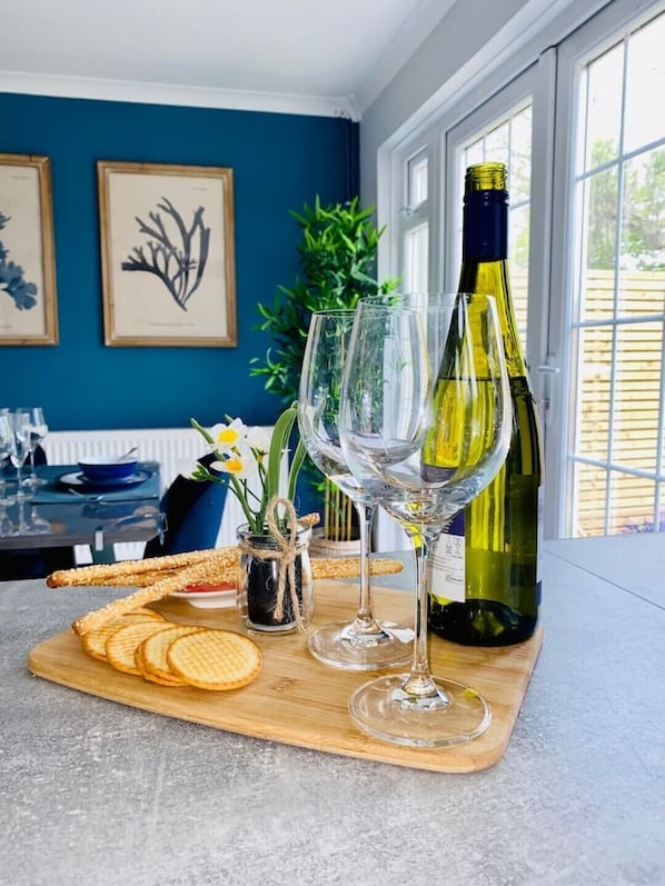 Une table à manger avec une bouteille de vin, deux verres à vin et une planche en bois sur laquelle sont disposés des crackers, ainsi qu’un petit vase avec des fleurs.
