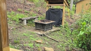 A wooden deck with a bench, a small fire pit, and a covered area in a forested setting.