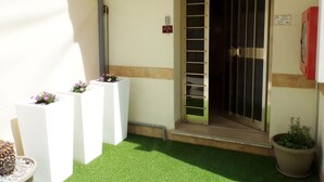 Entrada de un hotel con puerta de vidrio, un pequeño espacio al aire libre con macetas y un extintor rojo en la pared.