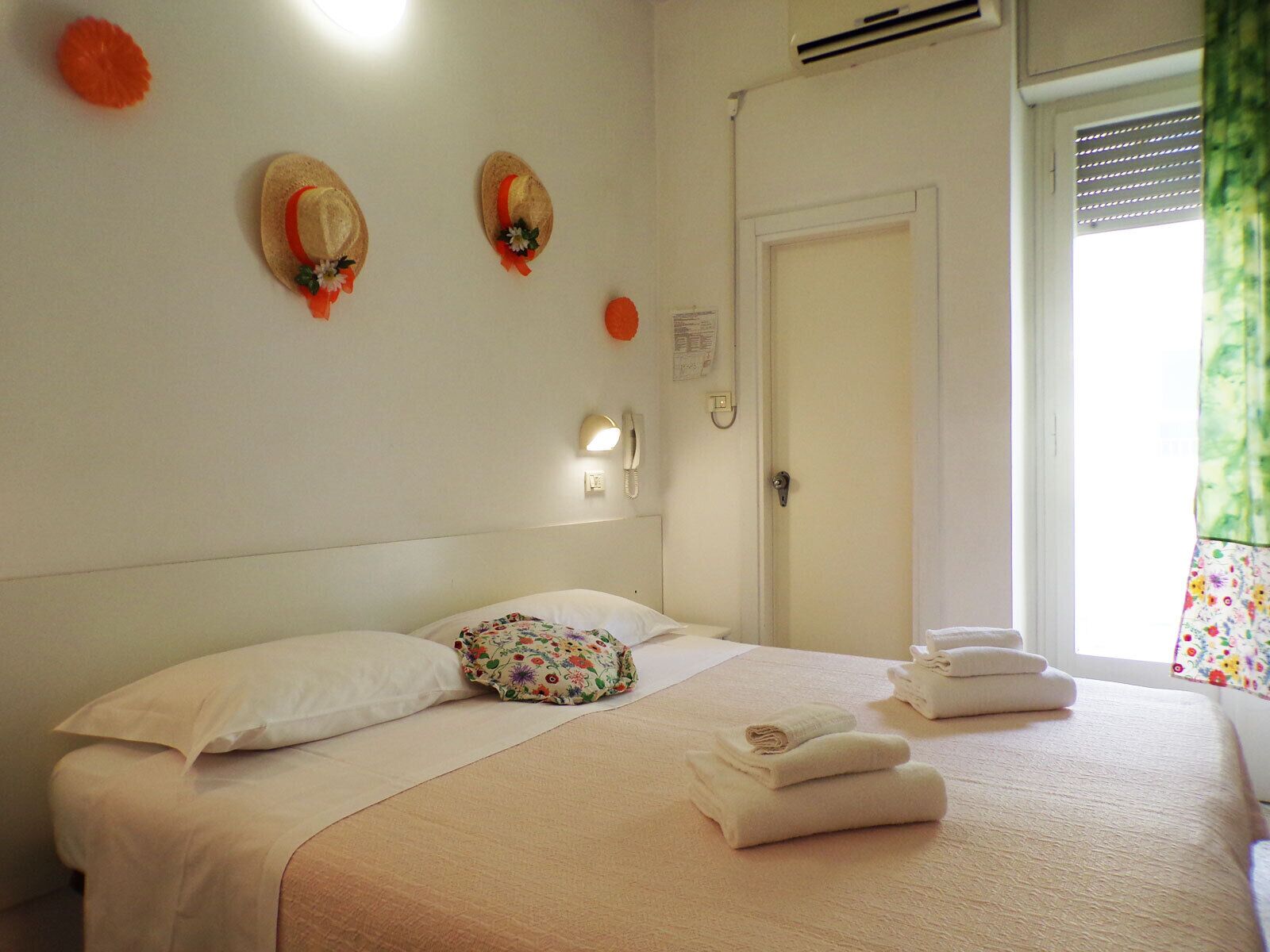 A neatly made bed with a floral pillow, two decorative hats on the wall, a bedside lamp, and a window with blinds.