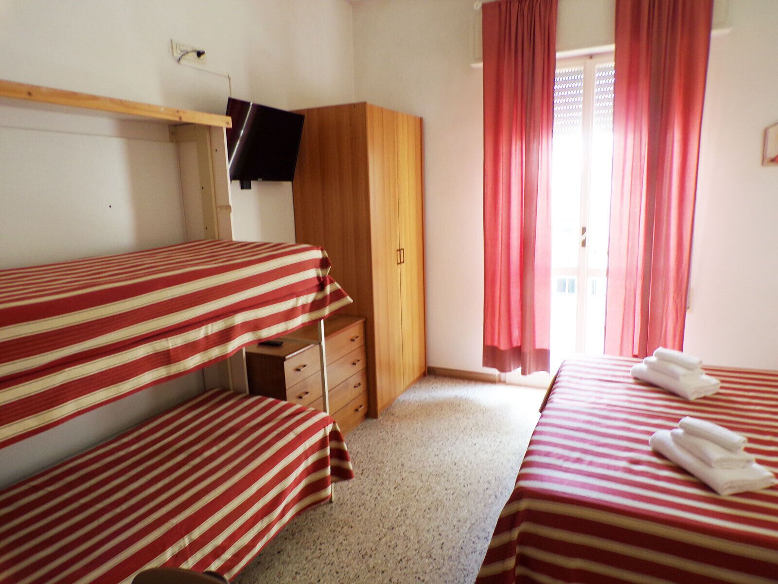 Quarto com duas camas, um guarda-roupa de madeira, televisão presa à parede e uma janela com cortinas vermelhas.