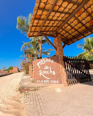 A sign with "Pousada do Rancho" and "Seja bem vindo" text, a brick wall, palm trees, and a paved area. - Pousada do Rancho (Barreirinhas)