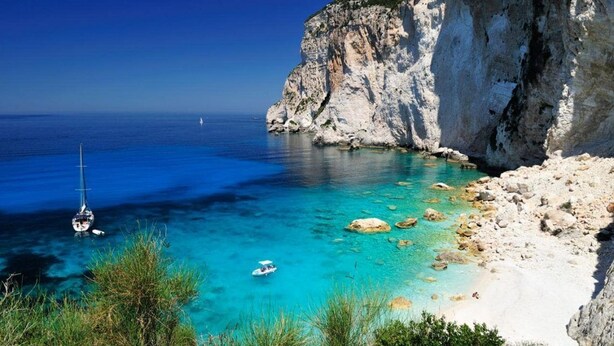 Un paysage côtier avec une eau turquoise et limpide, une côte rocheuse et un voilier.