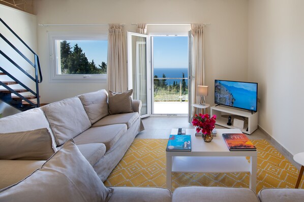 A living room with a sofa, a coffee table with books and flowers, a TV, and a balcony with a view of the sea.