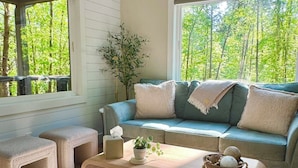 Ein gemütliches Wohnzimmer mit einer Couch, Hocker, einem Couchtisch und einem großen Fenster mit Blick auf Bäume.