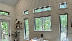 A spacious living room with a brown leather sofa, a coffee table, and a ceiling fan. There are multiple windows providing natural light and views of the outdoors. - The Stone - Modern Cabin on 40+ acres, 10 mins from Tryon Equestrian Center TIEC (Rutherfordton)