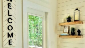 A modern interior with a 'WELCOME' sign, a glass door leading to a balcony, and a shelf with decorative items. - The Stone - Modern Cabin on 40+ acres, 10 mins from Tryon Equestrian Center TIEC (Rutherfordton)
