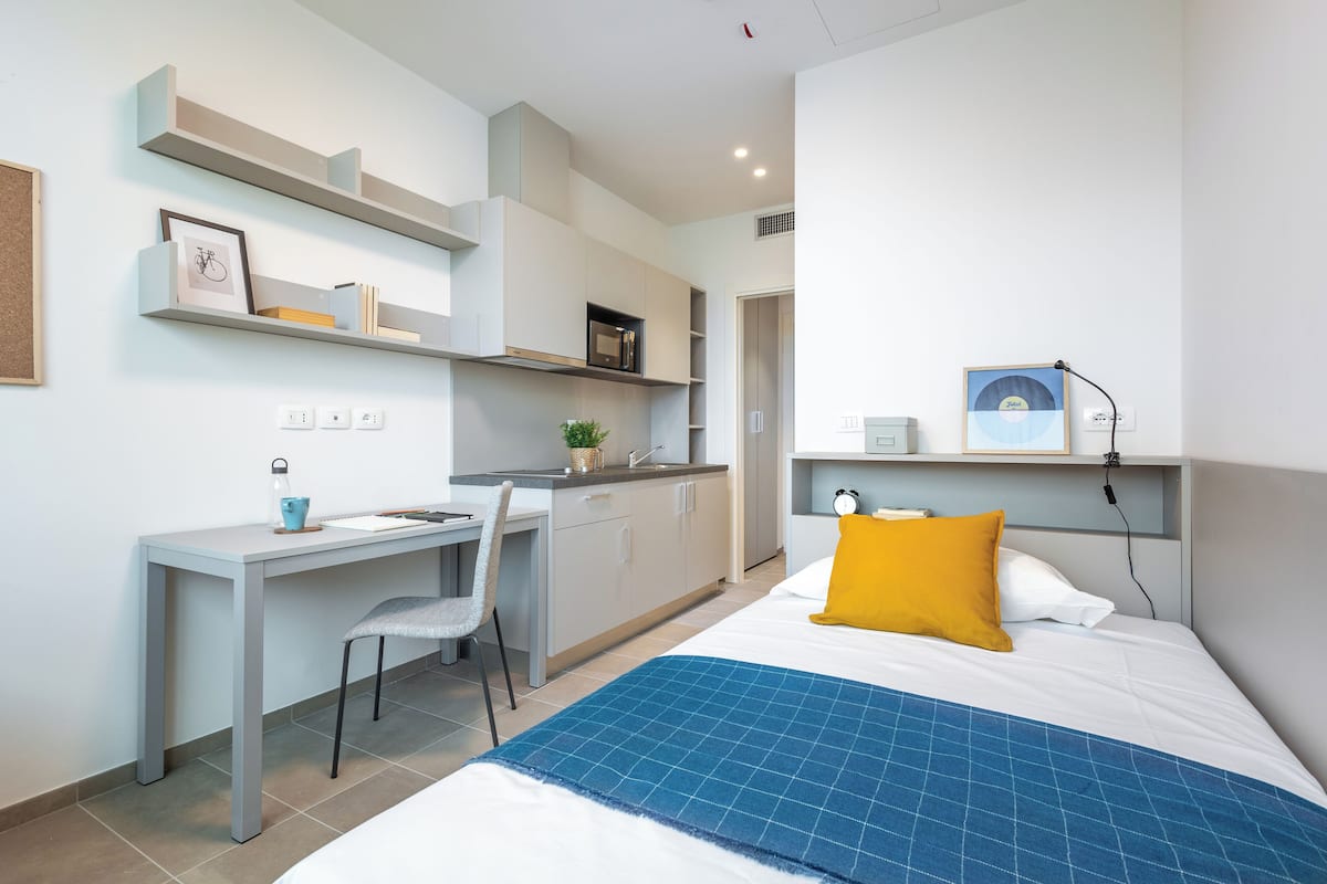 A modern bedroom with a bed, a desk, and a chair. There are shelves above the bed and a small kitchenette area.