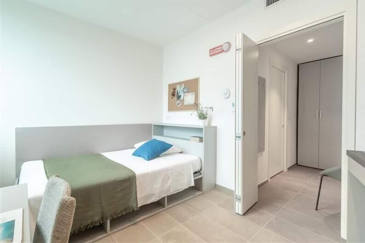 A small, neatly organized room with a bed, a desk, and a wardrobe.