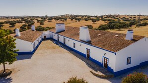 A white building with blue trim and multiple chimneys, surrounded by a gravel courtyard and a rural landscape. - Herdade dos Mestres (Alandroal)