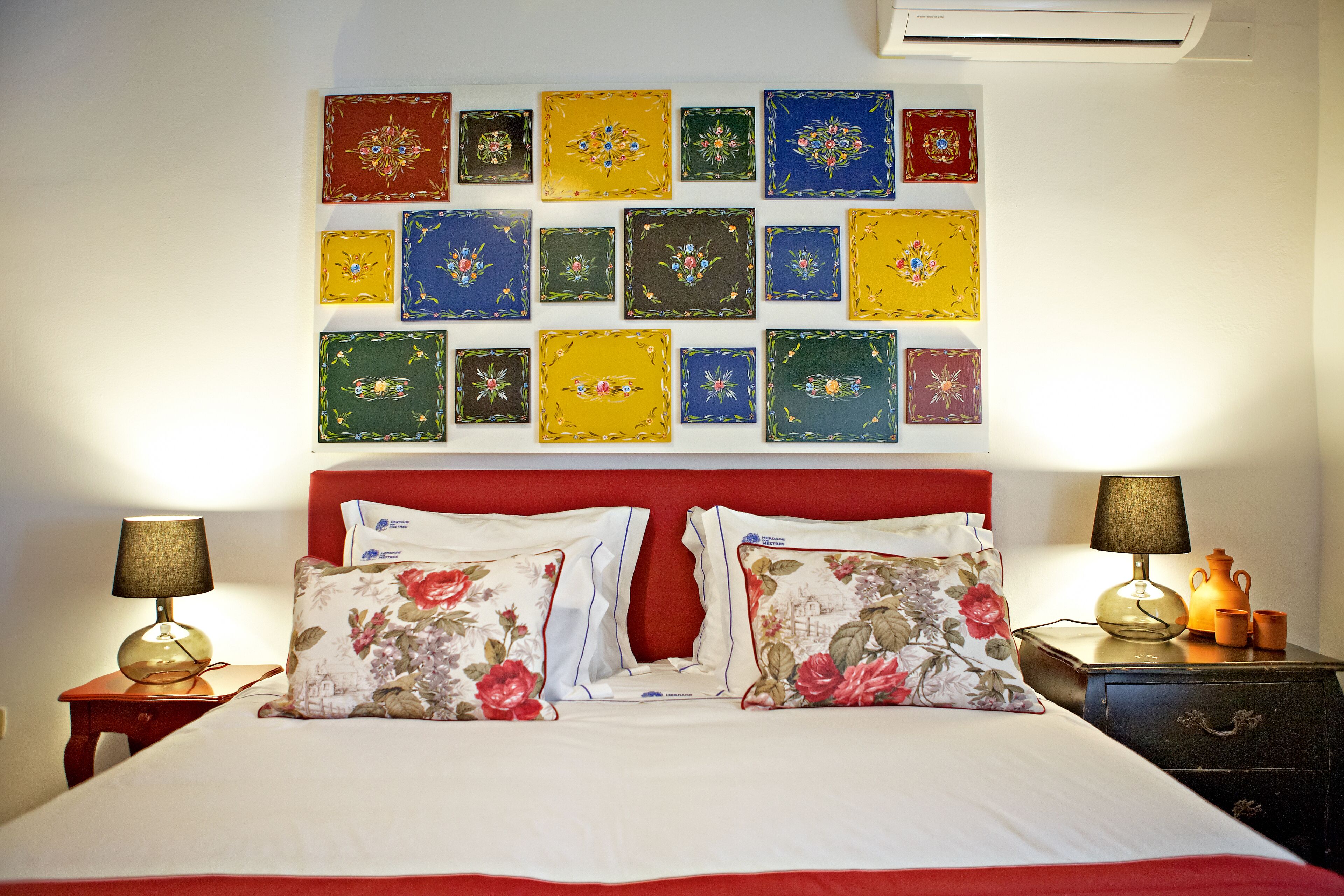 Un dormitorio ordenado con cabecera roja, almohadas estampadas con flores y una pared decorada con coloridas obras de arte cuadradas.