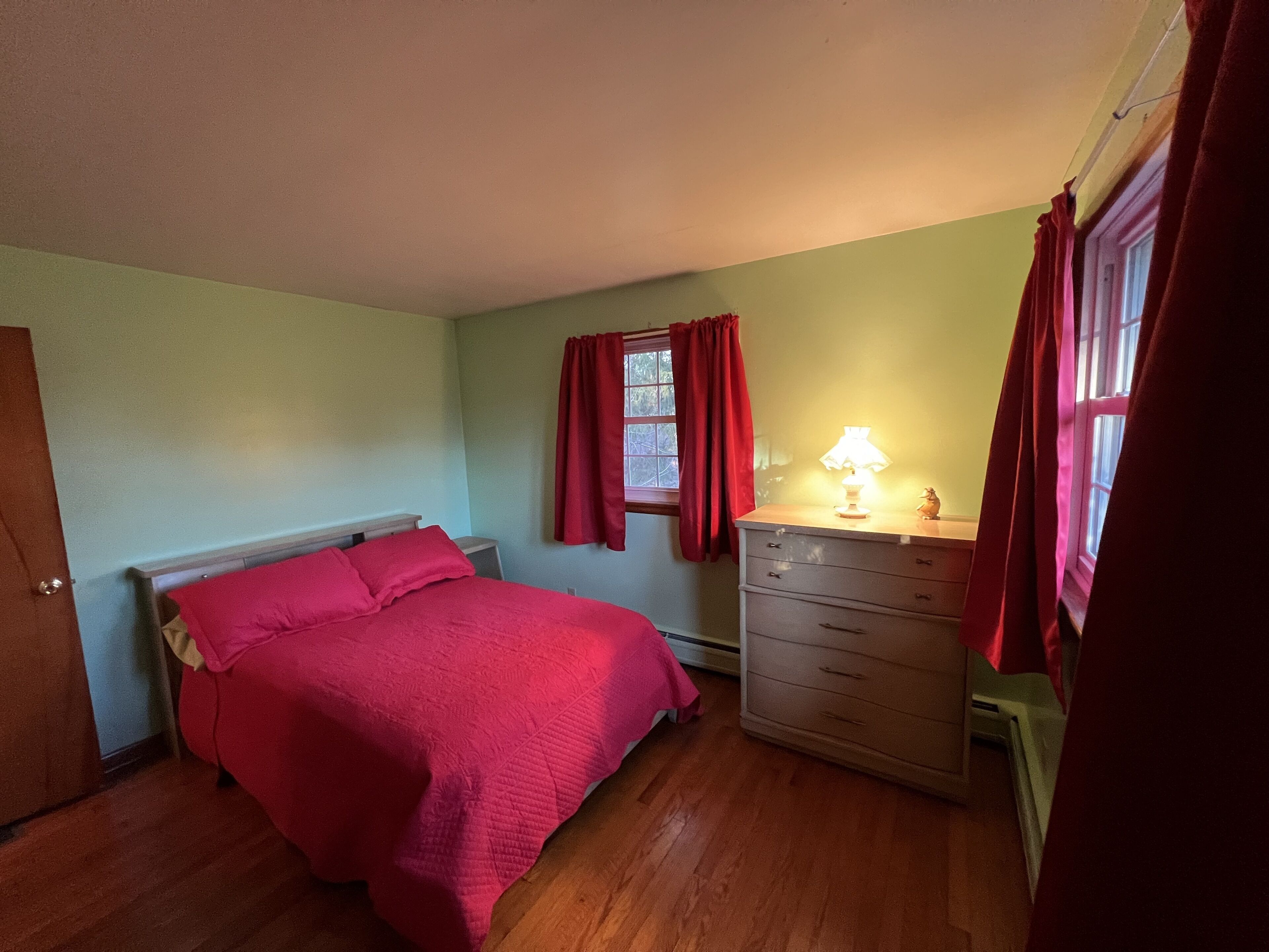 A bedroom with a bed, a dresser, a window with curtains, and a lamp.