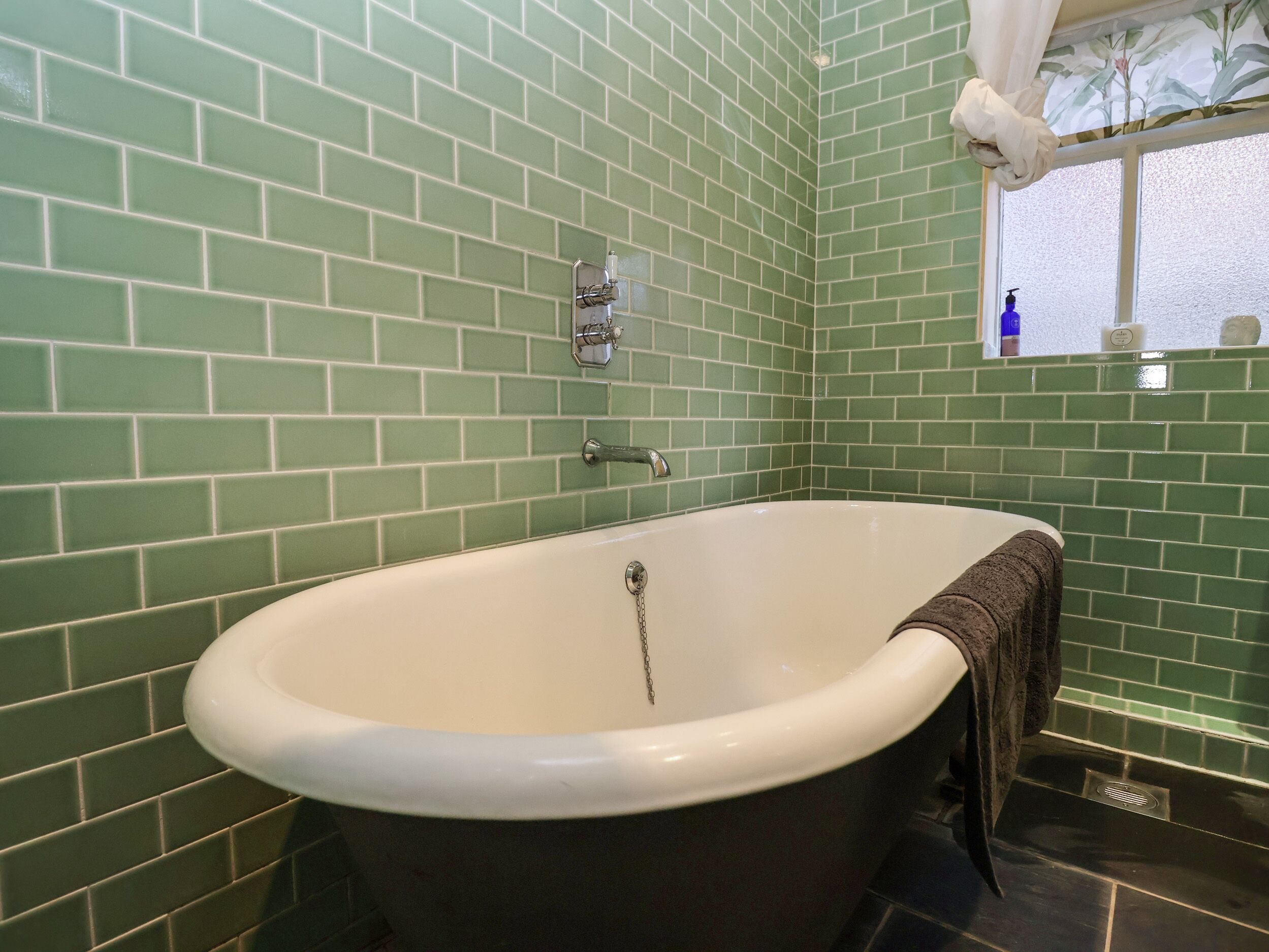 A bathroom with green tiled walls, a white bathtub, a towel hanging on the tub, a window with a view, and a blue bottle on the windowsill.
