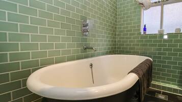 A bathroom with green tiled walls, a white bathtub, a towel hanging on the tub, a window with a view, and a blue bottle on the windowsill.