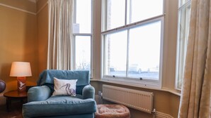 A room with a blue armchair, a small round ottoman, a lamp, and a view of the outside through large windows. - 11a Market Cross Place (Aldeburgh)