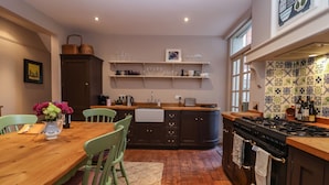 A kitchen with wooden cabinets, a dining table with chairs, a stove, and a window with patterned tiles. - 11a Market Cross Place (Aldeburgh)