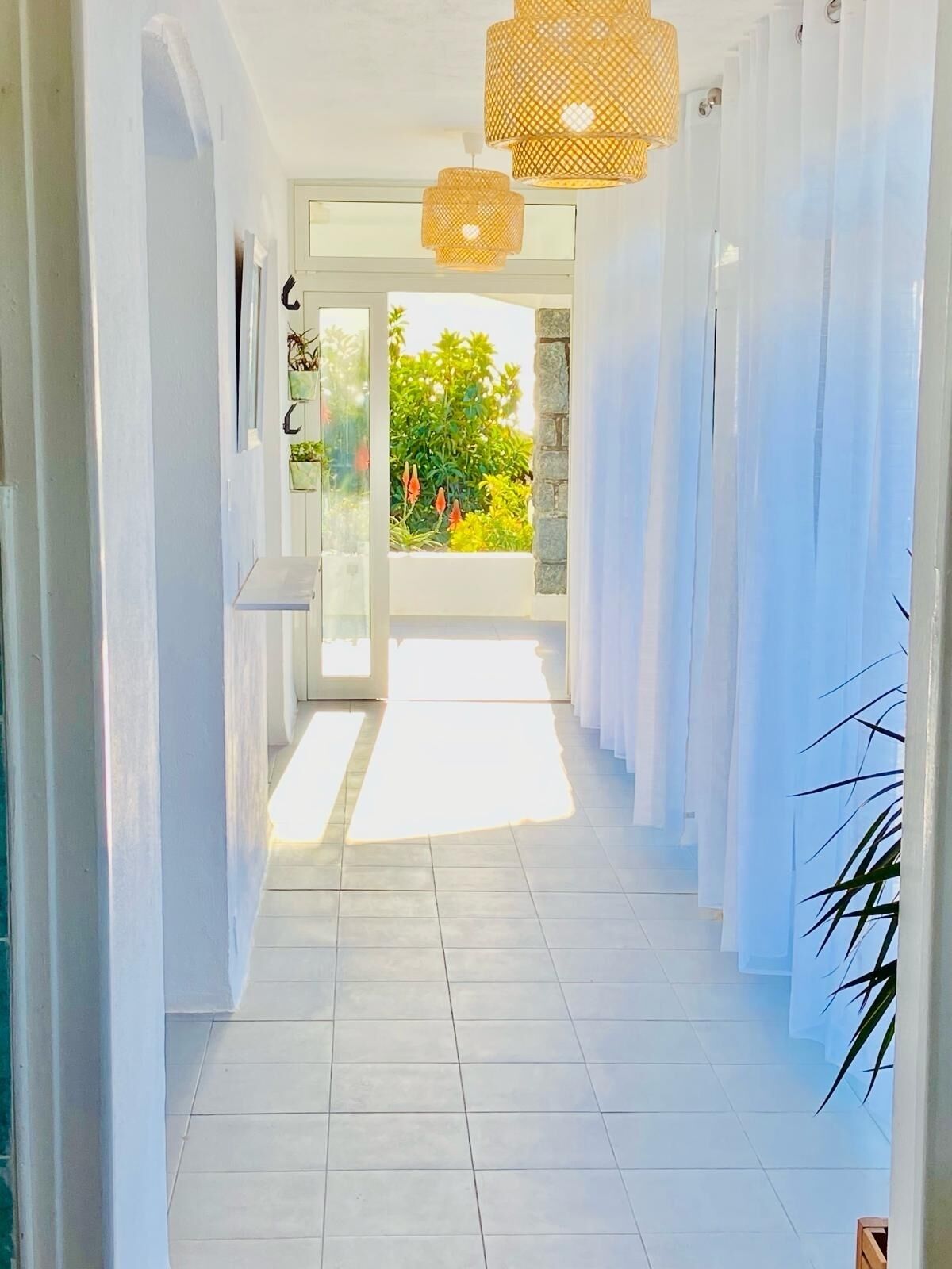 A bright hallway with white walls, a tiled floor, and a glass door leading to a balcony with plants.