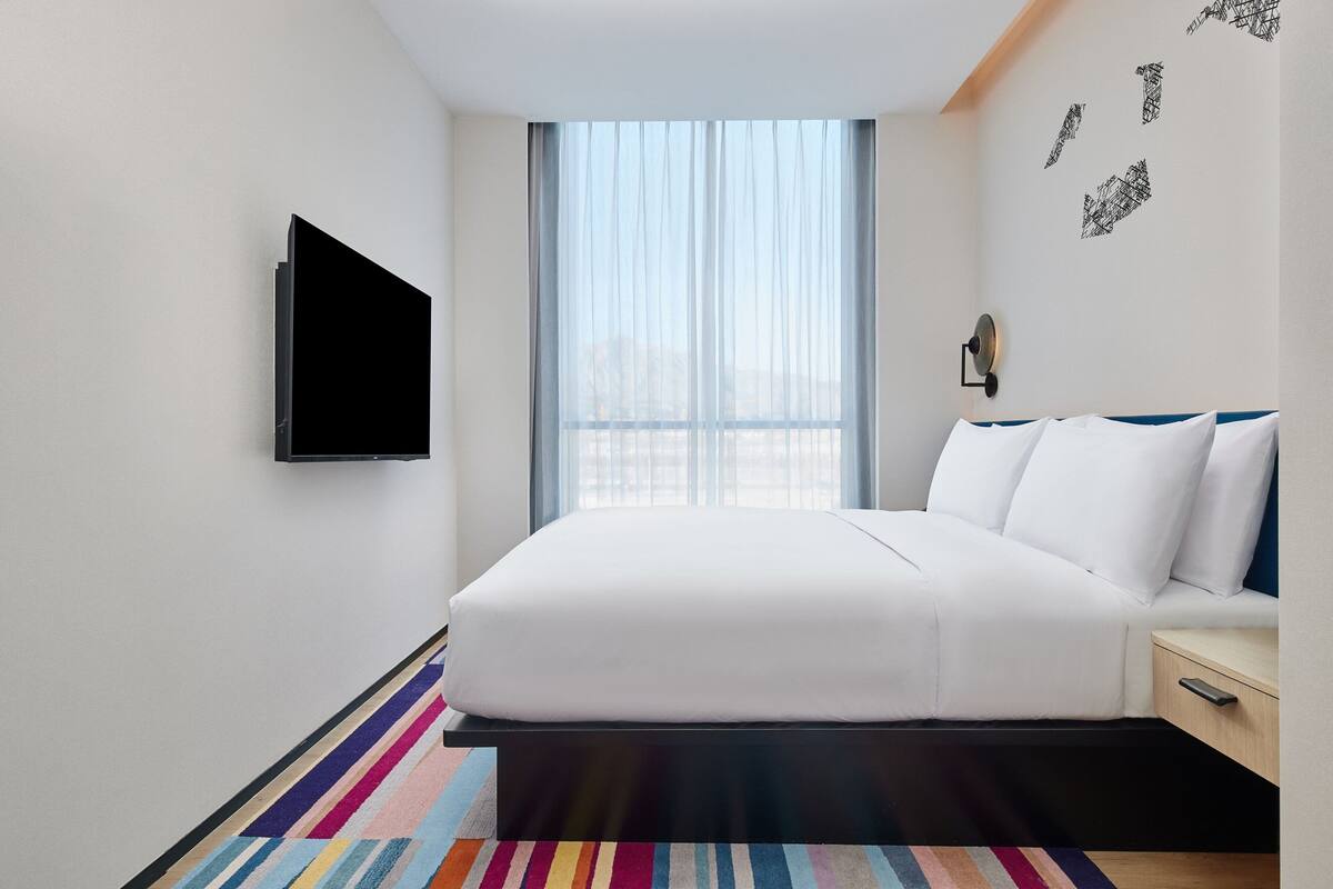 A modern hotel room with a large bed, a flat-screen TV, a bedside table, and a window with sheer curtains.