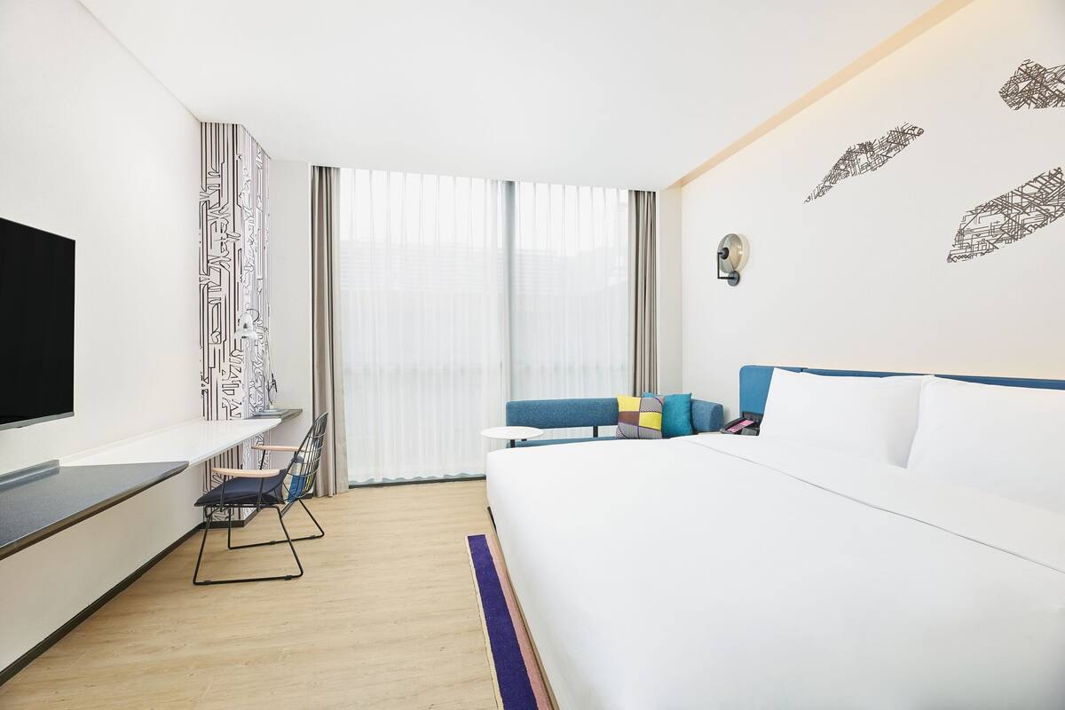 A modern hotel room with a large bed, a flat-screen TV, a desk, and a window with curtains.