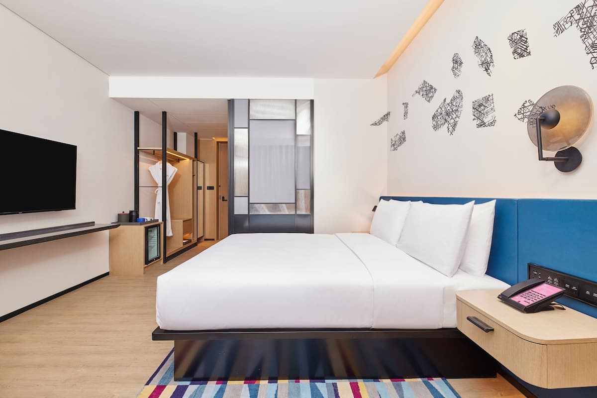 A modern hotel room with a large bed, a flat-screen TV, a bedside table with a phone, and a view of the hallway.
