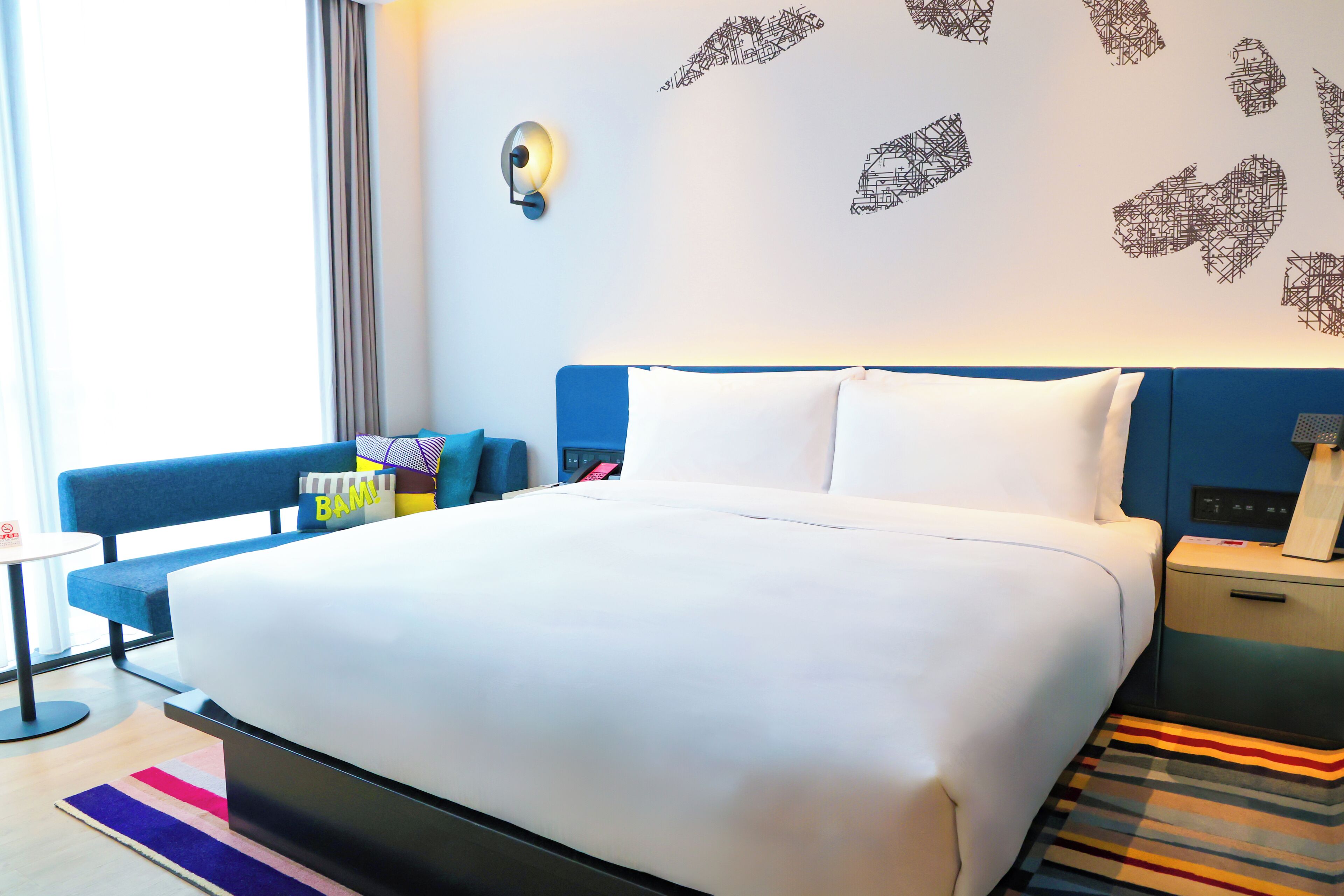 Ein modernes Hotelzimmer mit einem großen Bett, einem blauen Kopfteil, einem Nachttisch mit Lampe und einem farbenfrohen StreifenTeppich.