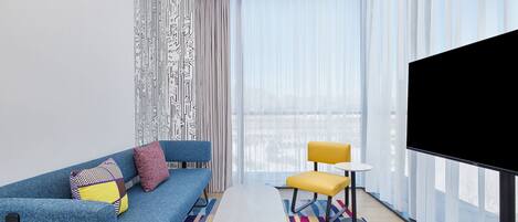 A modern hotel room with a blue sofa, a small table, a yellow chair, and a large TV screen.