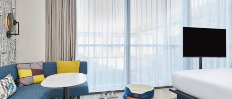 A hotel room with a sofa, a small round table, a pet bed, a TV, and a window with sheer curtains.