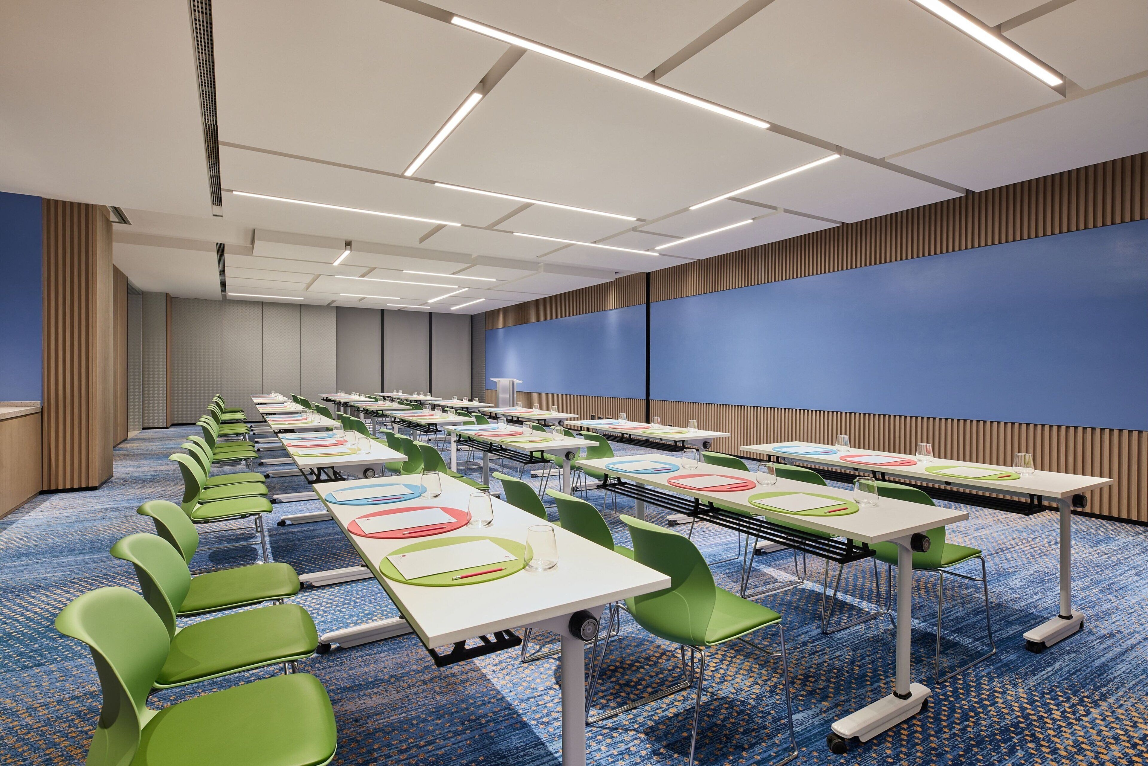 A conference room with rows of tables and chairs, a blue carpet, and a large screen wall.