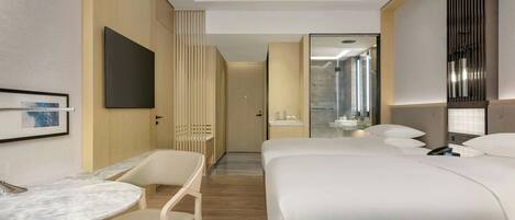 A hotel room with a bed, a desk with a chair, a TV, and a bathroom visible through a glass door.